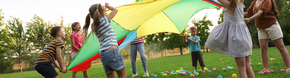 © iStock.com/Liudmila Chernetska Gruppe von Kindern und Lehrern, die mit Regenbogenspielplatz Fallschirm auf grünem Gras spielen. Sommercamp-Aktivität
