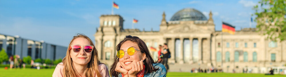 © iStock.com/frantic00 Zwei junge glückliche Mädchen, die eine Sonnenbrille tragen, die auf einem Gras liegt und vor dem Bundestagsgebäude in Berlin ihren Spaß haben. Auslandsstudium und Reisen in Deutschland Konzept
