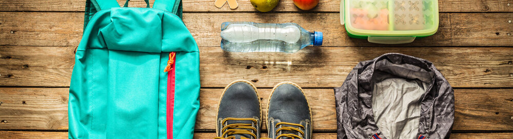 © iStock.com/Cleardesign1 Reisen - Packen (Vorbereiten) für das Konzept der Abenteuerschulausflug. Rucksack, Stiefel, Jacke, Lunchbox, Wasser und Obst auf Holzhintergrund von oben aufgenommen (Flat Lay).